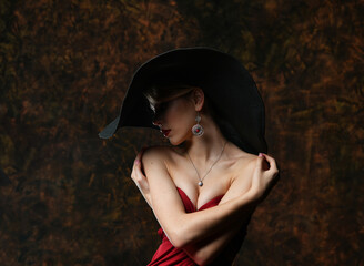 beautiful girl in a wide-brimmed hat and in a burgundy evening dress hugs herself on a black background