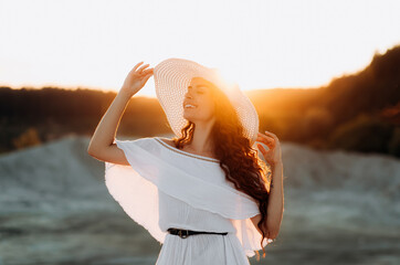 Pretty girl with wavy long hair in a white dress and hat at sunset, vacation concept.