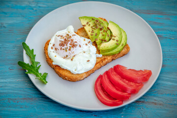 Toast with poached egg, flax seeds, avocado and tomato slices, arugula leaves on a plate. Healthy meal for breakfast.