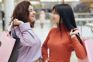 Two happy girlfriends looking smiling at each other while standing with shopping bags in mall, ladies wearing casual attire, being in good mood, enjoying of buying new clothing.