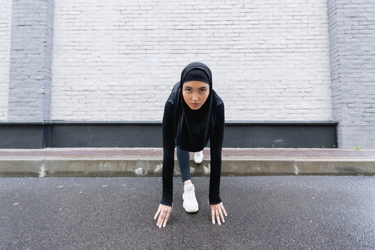 Young Muslim sportswoman in hijab and sportswear exercising near brick wall