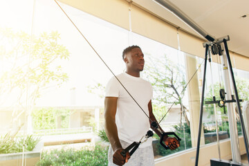 Muscular African American sportsman doing cable fly with exercise machine standing against window...