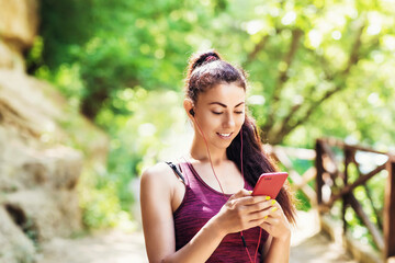Fototapeta premium Beautiful girl athlete chooses a track in a smartphone. Portrait of a beautiful sports model in the park with a phone in his hand