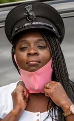 England, UK. 2020.   Portrait of a woman chauffeur with uniform cap and braided hair adjusting her pink coloured facemask during the Covid-19 outbreak
