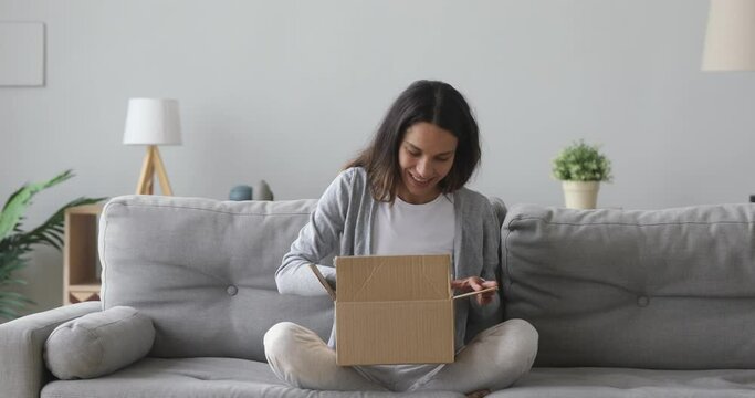 Happy young woman customer sit on sofa open parcel carton box satisfied with online shop order delivery at home, smiling girl consumer unpack package receive good purchase by postal shipping concept