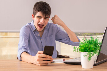 young male teenager or student at the desk with cellphone and computer