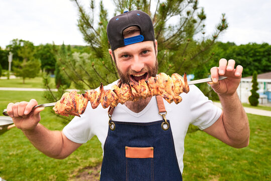 A young man with grilled chicken wings on a skewer in the backyard of a cottage.