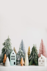 Christmas little houses and trees on white background. Festive modern decor. Happy holidays. Miniature cozy village, ceramic houses, wooden and handmade christmas trees. Space for text
