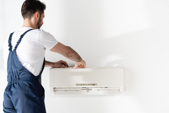 Repairman In Overalls Fixing Air Conditioner On White Wall