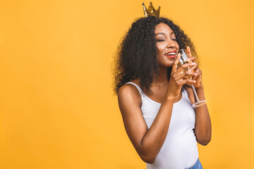 Happy birthday! Portrait of happy African-American black woman with glass of champagne and golden crown isolated over yellow background.