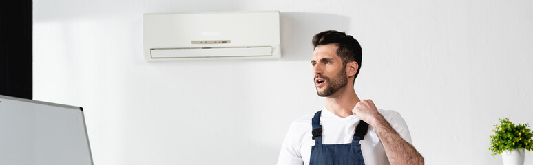 panoramic crop of repairman standing with toolbox near broken air conditioner and suffering from heat