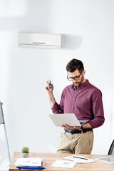 young businessman holding laptop and switching on air conditioner with remote controller