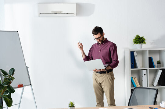 handsome businessman holding laptop and switching on air conditioner with remote controller