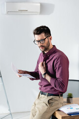 Fototapeta premium displeased businessman holding paper and touching shirt while suffering from heat in office