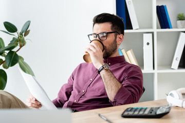selective focus of young businessman drinking coffee to go while studying document at workplace