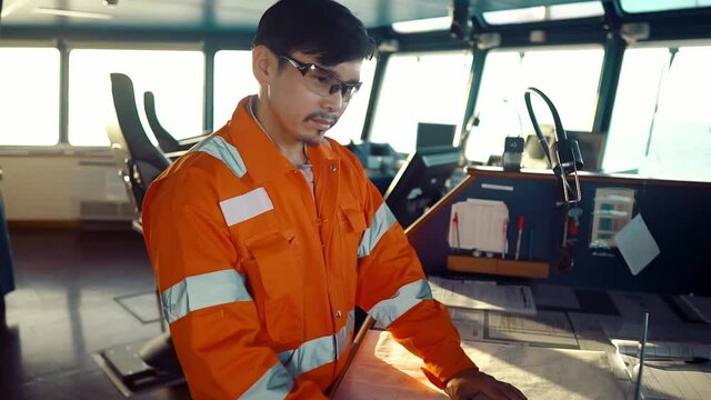 Filipino deck Officer on bridge of vessel or ship wearing coverall during navigaton watch at sea . He is using laptop, electronic paperwork at sea, concept of reporting
