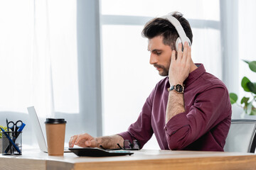 concentrated businessman in wireless headphones typing on laptop near coffee to go
