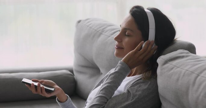 Calm happy young woman teen girl wear wireless headphones holding smart phone relaxing with eyes closed listening mobile music on smartphone in player app feel no stress free sit on sofa at home