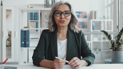 Beautiful senior businesswoman in formalwear and glasses sitting at desk in the office, looking at camera and having discussion while video calling during workd - Powered by Adobe