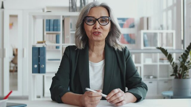 Beautiful senior businesswoman in formalwear and glasses sitting at desk in the office, looking at camera and having discussion while video calling during workd