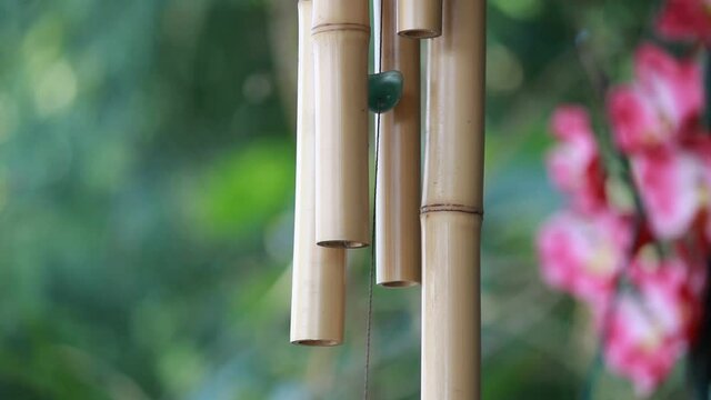 Sinos de vento feitos de bambu se mexendo em um a brisa num jardim com flores ao fundo