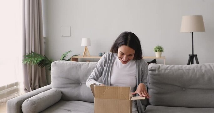 Excited girl customer open cardboard box feel satisfied sit on sofa, overjoyed young woman consumer get gift in postal parcel unpack package at home happy about fast shipment online purchase delivery