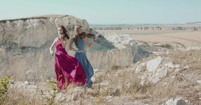 Female duet in elegant blowing dresses plays the violin among picturesque rocks