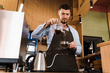 Pouring coffee in glass pot in cafe by handsome bearded barista. Coffee brewing syphon and...