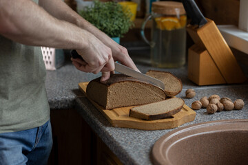Man's hand cutting rye bread on wooden board with knife on rustic style kitchen. Set of knives and some walnuts on table. Traditional nutrition concept. Vitamin B6 B12 Natural Source