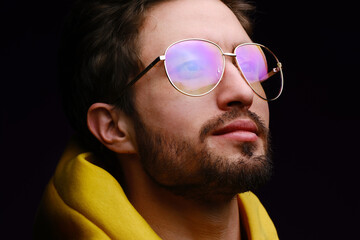 A young man of 25-30 years in glasses and a yellow sweatshirt emotionally poses on a black background. 