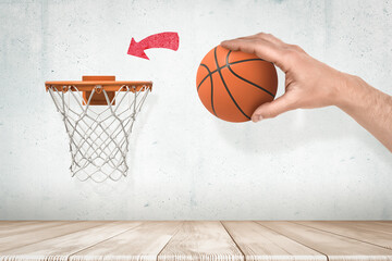 Man's hand holding orange basketball ready to throw it into basketball hoop fixed on grungy white wall with red painted arrow pointing at goal.