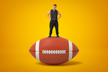 Tiny athletic man in black tracksuit standing on brown oval ball for American football on amber background.