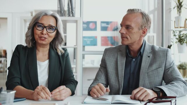 Beautiful senior businesswoman and professional middle-aged entrepreneur sitting together at desk in office and speaking at camera while having meeting via online video call