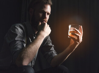 A handsome young man with a beard is sitting in a dark room with a glass of alcohol in his hands. The concept of rest and relaxation, booze and loneliness