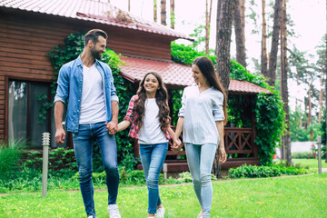 Obraz premium Young happy modern parents with a cute daughter are walking outdoors and holding hands together on suburban house background.