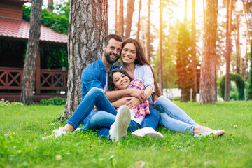 Fototapeta premium Beautiful happy family while sitting together on the grass and hugging each other, relaxing outdoors on suburban house background.
