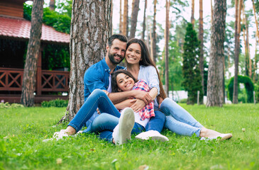 Fototapeta premium Beautiful happy family while sitting together on the grass and hugging each other, relaxing outdoors on suburban house background.
