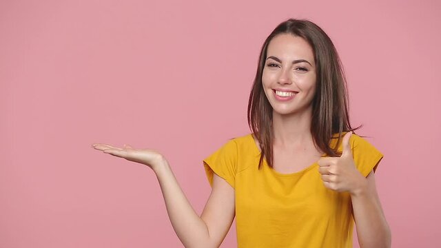 Cute young woman 20s years old in yellow t-shirt posing look camera pointing fingers hands on empty palm copy space aside workspace isolated on pastel pink background studio. People lifestyle concept