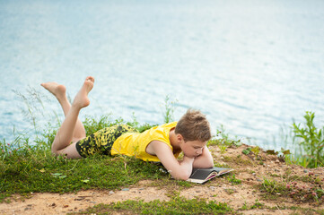 A boy alone lies on the river bank on a sunny summer day and reads a book. The teenager is resting alone.