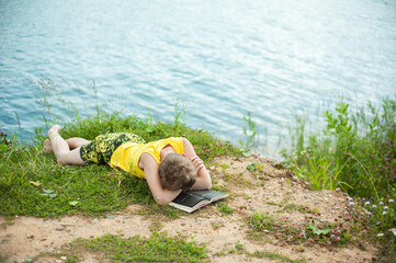 A teenager lies on the banks of a river or sea with a book. He fell asleep. The boy rests alone on a hot sunny day.