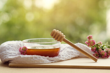 Honey dripping from a dipper in a glass bowl. Close-up. Healthy organic golden honey with a wooden spoon of honey, flowers of clover and white towels on a wooden table on a sunny day. Copyspace