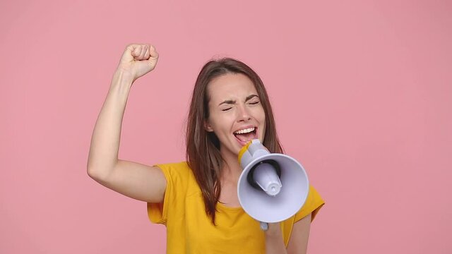Angry protesting young girl 20s years old in yellow t-shirt posing isolated on pastel pink background studio. People strikes pickets lifestyle concept. Look camera scream in megaphone say i am woman