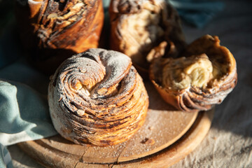 Homemade cruffin breads with dried fruits, cocoa and cinnamon fillings on wooden board and blooming cherry twig on linen tablecloth .