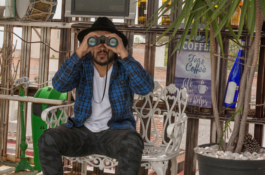 Hombre joven con camisa a cuadros y sombrero leyendo sobre la terraza, y usando binoculares 