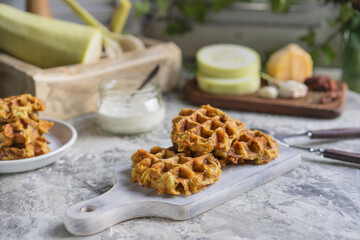 Savory squash or zucchi waffles with cheese, herbs and white sauce. Homemade baked waffles on grey textured table at kitchen, natural light, organic products on background. Selective focus, copy space