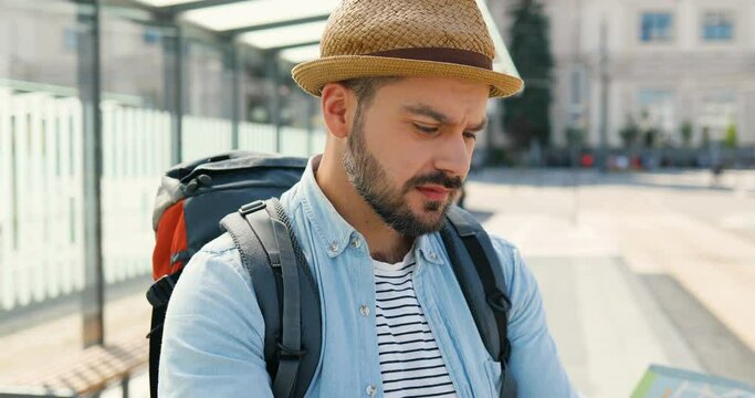 Young handsome Caucasian man tourist in hat with backpack holding city map and planning route. Looking for way in downtown train station. Attractive male traveller holding plan of town at bus stop.