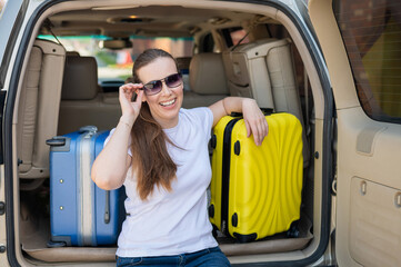 European woman in sunglasses packs her bags for a car trip. Beautiful happy girl sitting in the trunk of a car and ready for travel. Yellow suitcase with things for summer vacation.