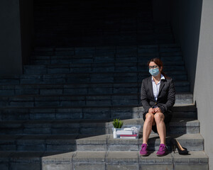 Unhappy woman in a mask sits with a box of personal stuff on the stairs outdoors. A female office employee was fired. The economic crisis at the height of the coronavirus epidemic.