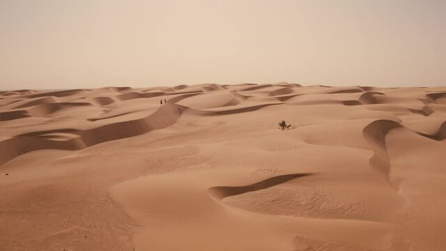 Aerial drone shot sweeping over perfect Sahara desert sand dunes as two people walk along the top of one of the dunes