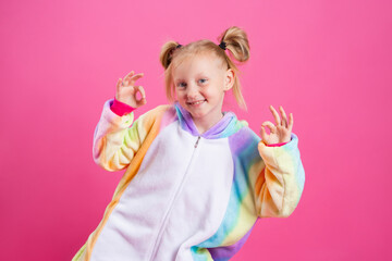 Happy little girl in kigurumi unicorn on a pink background rejoices in multi-colored confetti, space for text